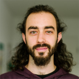 Alex has white skin, green eyes, a thick dark beard and dark long hair though with noticeable receding hairline.